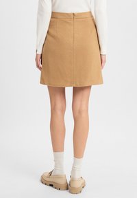 Beige Rock aus einem weichen Stoff, mit einer subtilen Textur, A-Linie Form und einem verdeckten Reißverschluss auf der Rückseite. Getragen mit beigen Schuhen und weißen Socken.