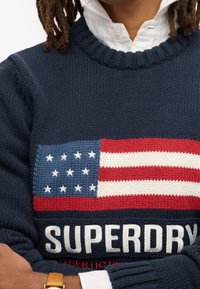 Pull en maille bleu marine mettant en avant un grand drapeau américain graphique aux couleurs rouge, blanc et bleu, avec le texte "SUPERDRY" en blanc en bas.