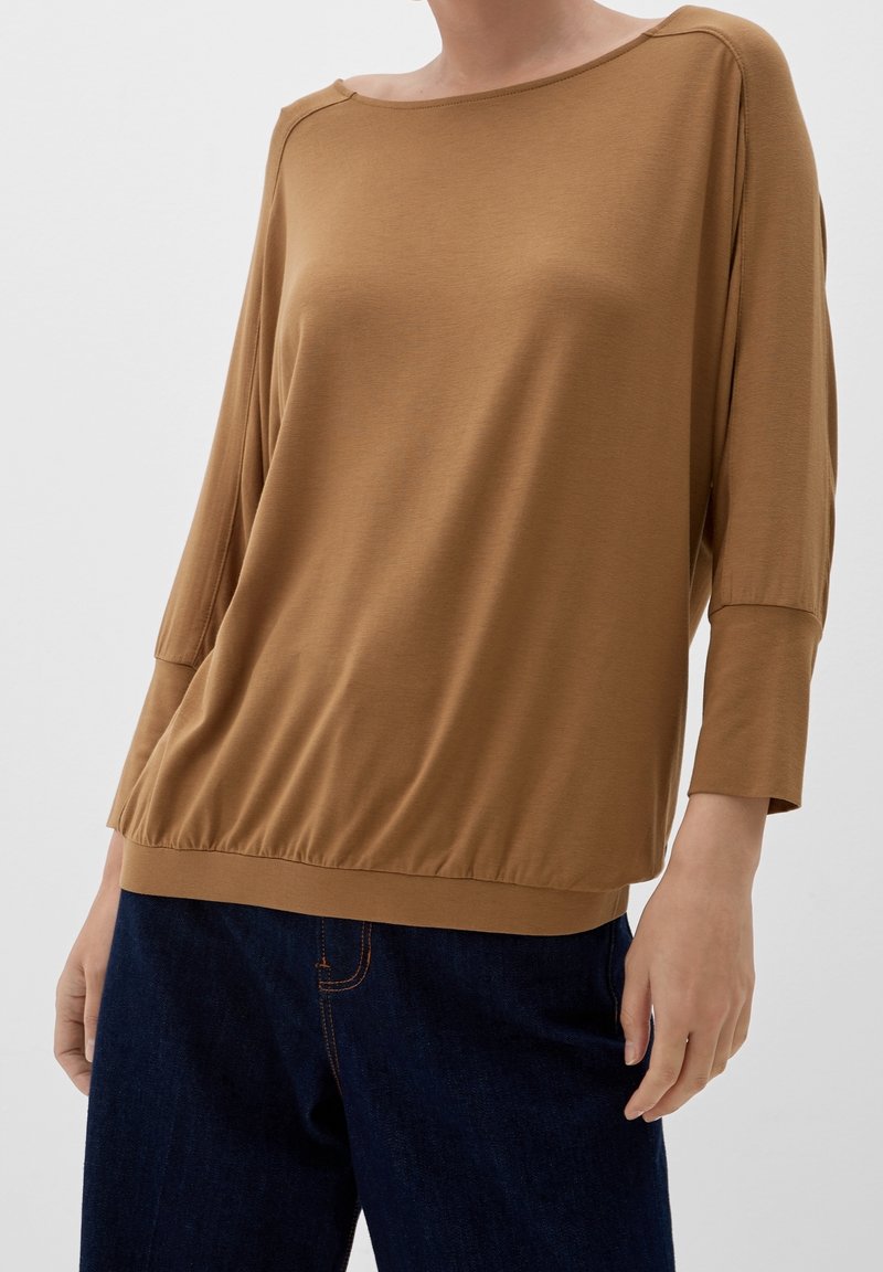 Brązowa, luźna bluzka z rękawami 3/4 i okrągłym dekoltem. Materiał jest gładki, z marszczonym dołem, zestawiona z ciemnymi dżinsami.