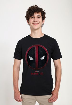 Ung mand med krøllet hår iført en sort Deadpool T-shirt med rød og sort logo og beige bukser, der smiler til kameraet.