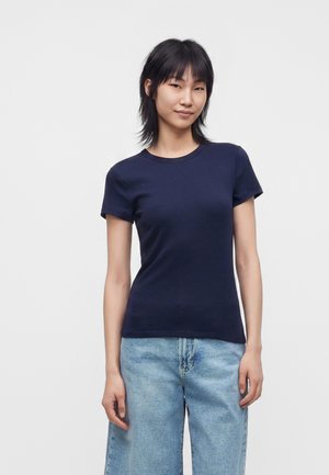 Jeune personne aux cheveux courts et noirs, portant un t-shirt bleu marine et un jean large bleu clair, debout devant un fond blanc uni.