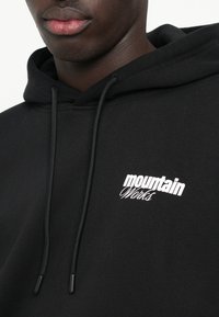 Czarna bluza z kapturem z miękkiego materiału, z białym logo "Mountain Works". Zawiera sznurki ze czarnymi końcówkami.