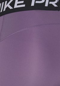 Fialové sportovní legíny s hladkou texturou, černý pás s bílým potiskem "Nike Pro". Viditelné detaily šití.