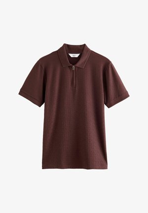 Ung mand med krøllet hår iført en kortærmet bordeaux polo med lynlås, der kigger let til siden mod en ensfarvet baggrund.