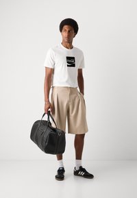 Witte T-shirt met zwart Coca-Cola logo, beige plooishorts, zwarte sporttas, zwart en blauwe sneakers, en witte sokken.