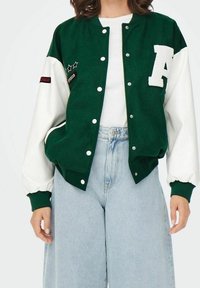 Zielona kurtka typu varsity z białymi rękawami, z haftowaną literą "A" i gwiazdkami. Wykonana z miękkiego materiału, z zapięciem na zatrzaski i bocznymi kieszeniami.
