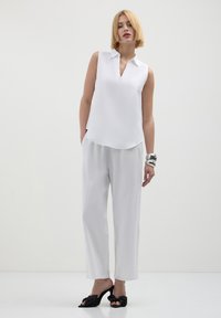 Donna con capelli biondi corti, che indossa una blusa senza maniche bianca, pantaloni bianchi, braccialetti argentati e sandali neri con tacco, in piedi davanti a uno sfondo bianco.