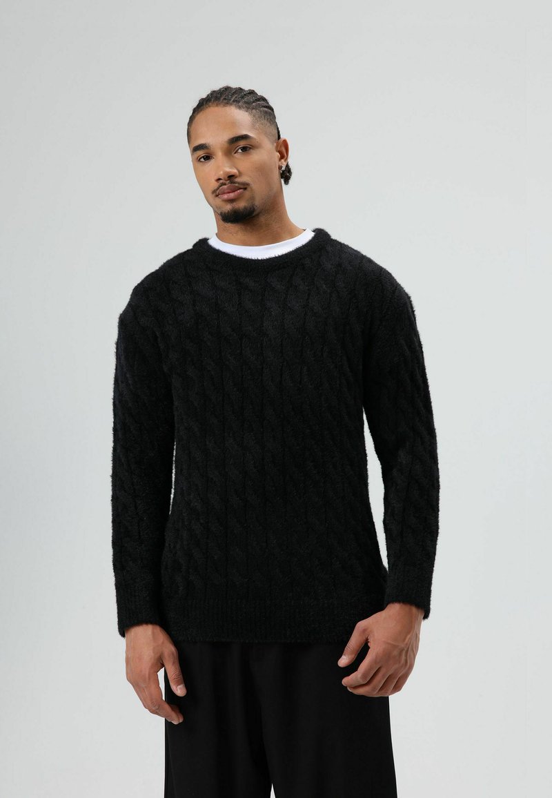Zwarte kabelgebreide trui met een gestructureerde afwerking, ronde halslijn en lange mouwen, gecombineerd met donkere broek. Zacht, comfortabel materiaal.