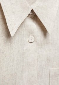 Camicia in lino beige chiaro con colletto a punta, dotata di chiusura a un bottone e taschino sul petto; dettagli in tessuto testurizzato visibili.