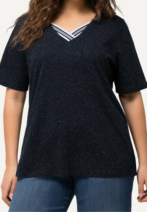 Vrouw die een zwart gespikkeld V-hals T-shirt draagt met witte gestreepte kraagdetail en blauwe jeans, staand tegen een effen achtergrond.