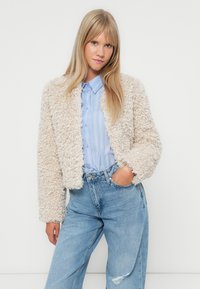 Giacca in fleece color crema, con design texturizzato e corto, indossata sopra una camicia a righe blu chiaro, abbinata a jeans blu a vita alta.