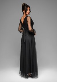 Abito da sera nero con maniche lunghe trasparenti, scollatura a V profondo sul retro e vita annodata. La gonna presenta strati di tulle per una silhouette fluida.