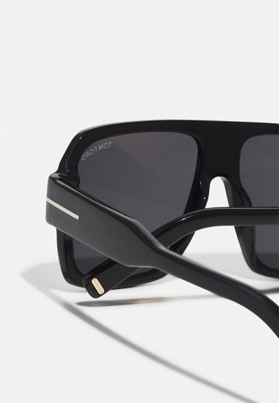 Occhiali da sole neri con design angolare. Presentano lenti scure e un accento in metallo dorato sulla montatura nera. Bracci testurizzati con dettagli del logo.