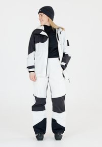 Skijack en -broek gemaakt van wit en zwart stof, met een modern ontwerp, meerdere zakken en een rits. Gebreide zwarte beanie.