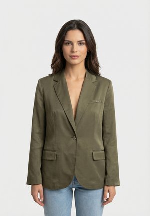 Donna che indossa un blazer verde oliva a bottone singolo e jeans azzurri, in piedi su uno sfondo chiaro e uniforme.