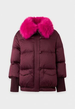 Femme portant une doudoune bordeaux avec un col en fourrure rose vif, un jean bleu et des baskets blanches, debout devant un fond blanc uni.