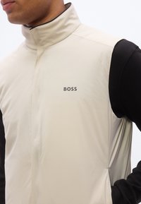 Κοντινό πλάνο ενός ατόμου που φοράει μπεζ αμάνικο γιλέκο με όρθιο γιακά και λογότυπο "BOSS" στο στήθος πάνω από μαύρο πουκάμισο με μακριά μανίκια.