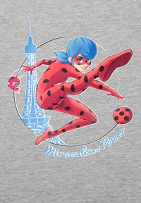 Roter Superheld mit schwarzen Punkten, blauen Haaren und einer Maske, bereit, einen Ball zu treten. Mit dem Eiffelturm und dem Text "Miraculous Power".