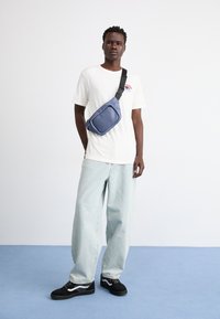 Blue nylon crossbody bag worn over a white t-shirt. Features a smooth texture, adjustable strap, and zippered compartment. Light-wash jeans and sneakers.