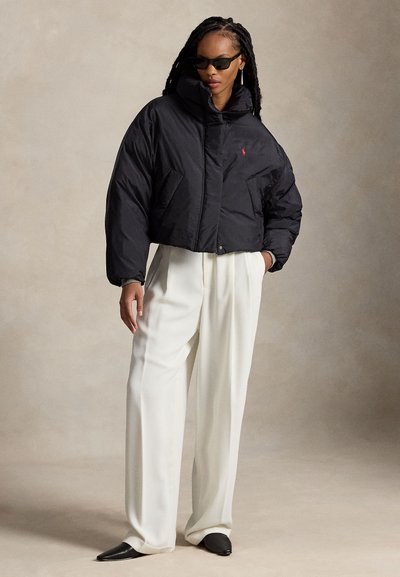 Femme aux longues tresses portant une veste matelassée noire courte, un pantalon blanc à jambes larges, des chaussures noires pointues et des lunettes de soleil noires, debout devant un fond uni.