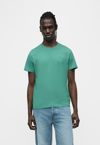 Πράσινο βαμβακερό μπλουζάκι με στρογγυλή λαιμόκοψη, κοντά μανίκια και μικρό λογότυπο τυπωμένο στο στήθος. Συνδυασμένο με ανοιχτό μπλε τζιν παντελόνι.