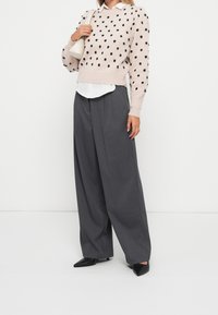 Maglione a maglia rosa chiaro con pois neri, abbinato a pantaloni grigi a gamba larga. La modella tiene una piccola borsa bianca e indossa tacchi neri.