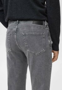 Man in een donkergrijze gewassen spijkerbroek en een zwart langearmshirt, met zicht van achteren van de taille tot halverwege het dijbeen.