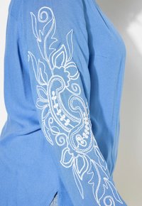 Blusa a maniche lunghe blu con ricami floreali bianchi lungo il braccio sinistro, realizzata in un tessuto morbido e leggero con una vestibilità comoda.