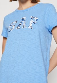 T-shirt bleu clair à manches courtes, arborant le logo "GAP" en imprimé floral. Texture douce et encolure ronde, coupe décontractée.