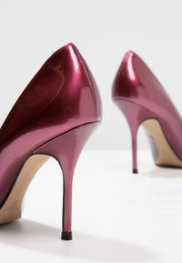 Paire de chaussures à talons hauts métalliques brillantes de couleur rose foncé avec des bouts pointus sur une surface blanche, vues de l'arrière.