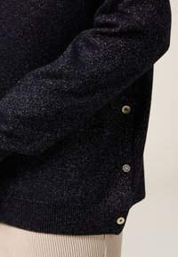 Granatowy sweter z metalicznym połyskiem, prążkowanym dołem i ozdobnymi guzikami. Posiada gładką, teksturowaną tkaninę oraz luźny krój.