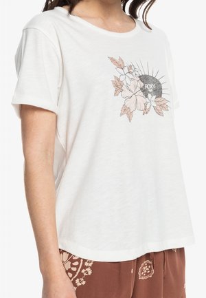 Chemise blanche à manches courtes avec un motif floral et le texte « ROXY » sur la poitrine, portée avec un pantalon marron à motifs.