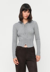 Joven con una sudadera de punto trenzado gris con cremallera y pantalones marrón oscuro, de pie contra un fondo blanco liso.