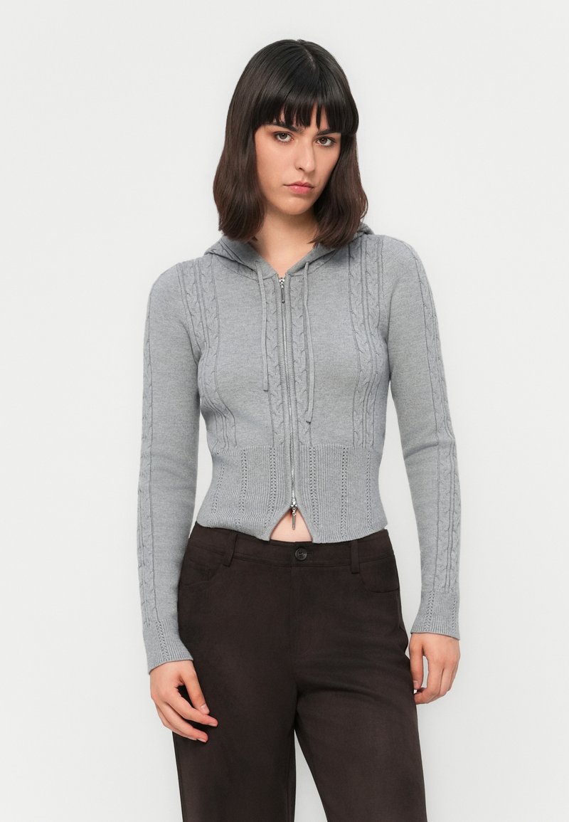 Młoda osoba w szarej bluzie z kapturem i zamkiem, o splocie warkoczowym, oraz ciemnobrązowych spodniach, stojąca na tle białego tła.