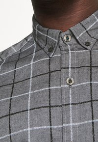 Camicia grigia con bottoni e motivo a quadri con linee nere e azzurro chiaro, colletto arrotondato e bottoni scuri. Tessuto con trama visibile.