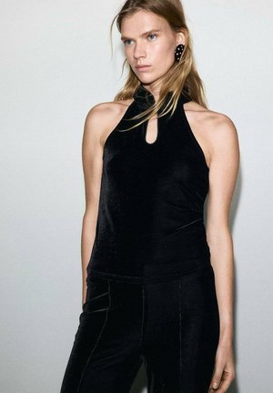 Top a halter in velluto nero con dettaglio a occhio di bottone. Texture liscia e design aderente, abbinato a pantaloni neri, creando un look monocromatico.