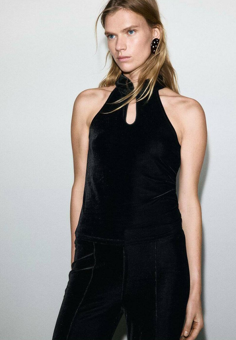Top a halter in velluto nero con dettaglio a occhio di bottone. Texture liscia e design aderente, abbinato a pantaloni neri, creando un look monocromatico.