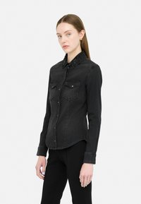 Camicia in denim nera con due tasche anteriori e colletto decorato con borchie. Presenta bottoni sul davanti e un orlo curvato.