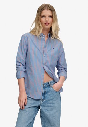 Mujer rubia con cabello ondulado que lleva una camisa abotonada a rayas azules y rosas y vaqueros azul claro, mano en el bolsillo, expresión neutral.