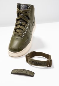 Vysoké olivově zelené tenisky s texturovanou kůží, krémovou podešví a dvěma odnímatelnými látkovými pásky označenými "FORCE" a "FEMALE" v černé barvě.