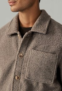 Braune, pelzige Jacke mit Kragen, großer aufgesetzter Tasche und Knopfverschluss. Die Textur wirkt weich und warm.