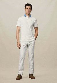 Valkoinen polo-paita vaaleansinisen kauluksen kanssa, slim-fit-muotoilu. Yhdistetty valkoisiin housuihin ja ruskeisiin kenkiin, esitetty neutraalia taustaa vasten.