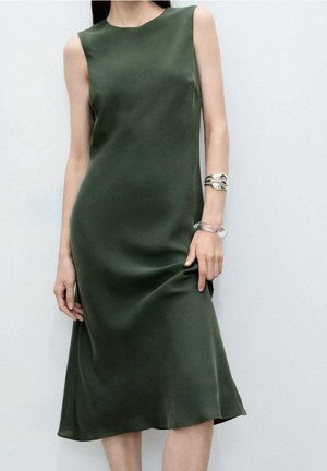 Kvinde iført ærmeløs midi-kjole i olivengrøn med glat stof, iført sølv- og klare armbånd på højre håndled mod en ensartet lys baggrund.