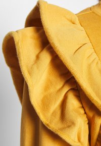 Žlutá rýškovaná tkanina s měkkou texturou. Má stažený design na rukávu, který zvýrazňuje hladký povrch a živou barvu.