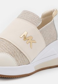 Sneaker beige senza lacci con texture intrecciata, fascia elastica, logo dorato MK, suola bianca e battistrada marrone.