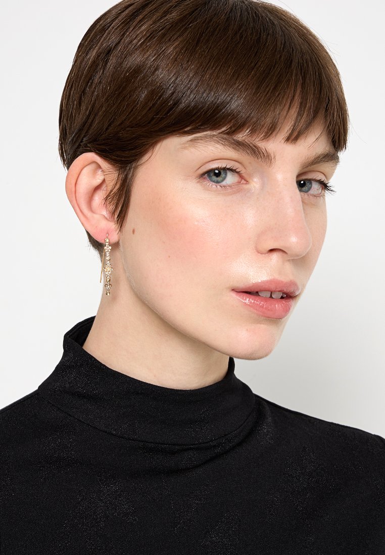 Goldfarbene hängende Ohrringe mit zarten Verzierungen, getragen von einem Modell in einem schwarzen Rollkragenpullover mit einem subtilen Schimmer.