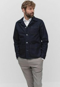 Mörk denimjacka med krage, framknappar och två påsydda fickor; kombinerad med ljusgrå byxor och en vit skjorta.