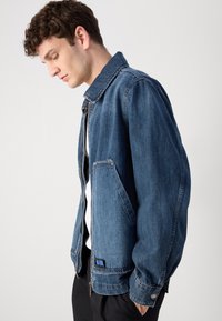 Jachetă din denim de culoare albastru închis, cu guler, închidere cu fermoar și buzunare laterale; are un material texturat și o etichetă de marcă vizibilă.