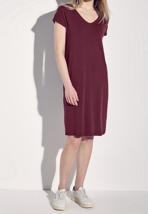 Femme portant une robe courte bordeaux à manches courtes, arrivant au genou, et des baskets blanches, debout devant un fond blanc uni.