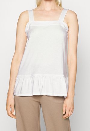 Biały bawełniany top na ramiączkach z kwadratowym dekoltem, szerokimi ramiączkami i falbanką na dole. Połączony z brązowymi spodniami.